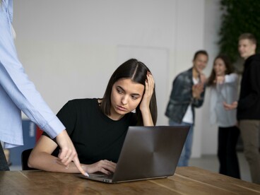 Un giovane donna, visibilmente turbata e con la testa appoggiata alla mano, è seduta davanti al laptop, mentre sullo sfondo altre persone la prendono in giro e ridono