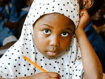 A young girl in Africa focuses on her writing during class, surrounded by classmates, capturing the spirit of learning and academic growth
