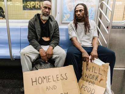 Two people sitting side by side in a subway, holding a handwritten sign that states they are homeless, representing housing insecurity in everyday public spaces