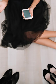 A woman with long legs sits on the floor of a stylish wardrobe, surrounded by an array of shoes. She’s wearing a chic black skirt and elegant silver high heels while using her smartphone to text and browse the internet
