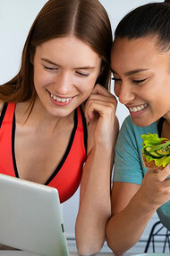 A woman in a kitchen, surrounded by healthy food, examines an online information source on the table