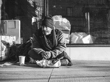 Framed by consumer waste and glass storefronts, the man’s presence critiques the contradiction of wealth and deprivation existing side by side