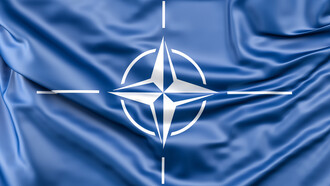 The NATO symbol resting on blue satin, conveying how global order is often staged as harmony while driven by strategic profit