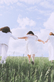 Ragazze di spalle corrono in una distesa di verde in abiti bianchi 