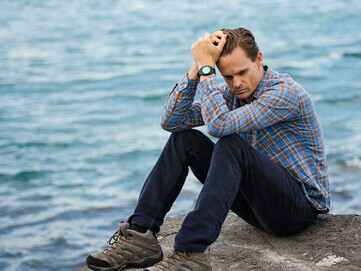 A man wearing a blue and brown plaid shirt touches his hair as he sits looking sad, depressed, and lonely