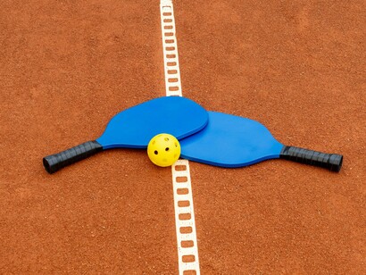 Two blue pickleball paddles and a bright yellow pickleball rest on a red court, illustrating the simple equipment needed for a sport that offers a fun, low-impact workout, improves hand-eye coordination, and provides a social outlet for all ages
