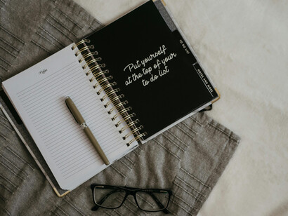 A pen rests on a notebook lying on a bed, its cover inscribed with the words ‘Put yourself at the top of your to do list'