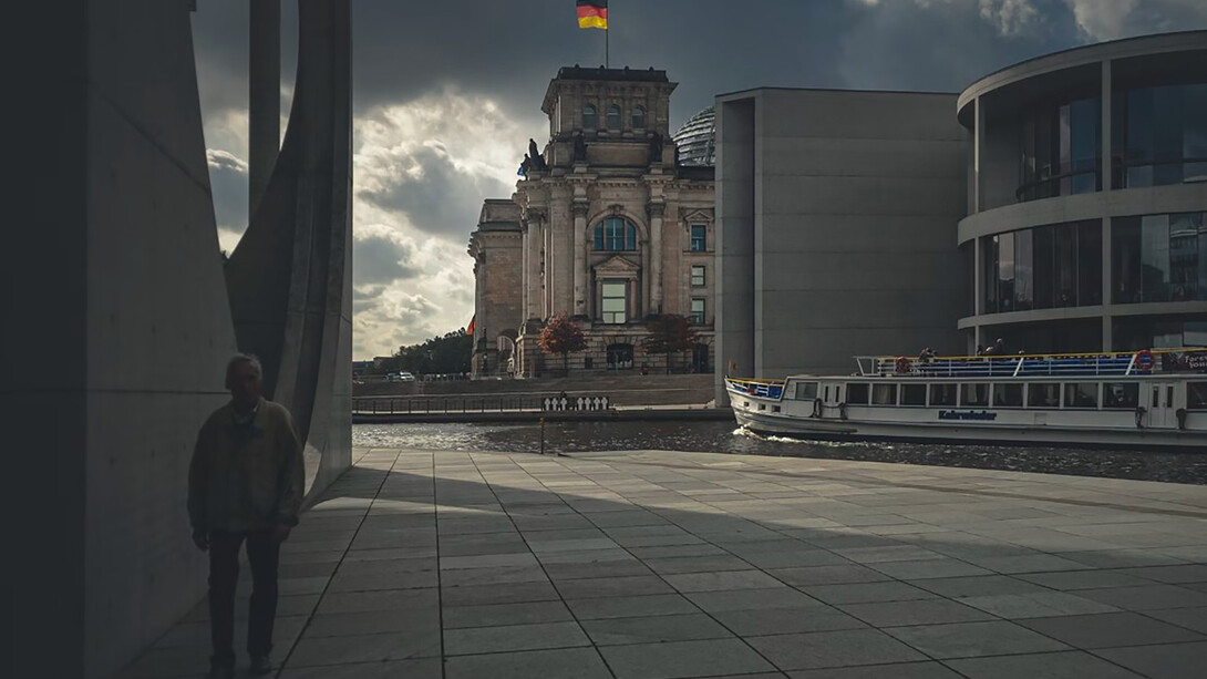 An elderly man walks along a gray concrete path beside a brown building in Berlin, Germany