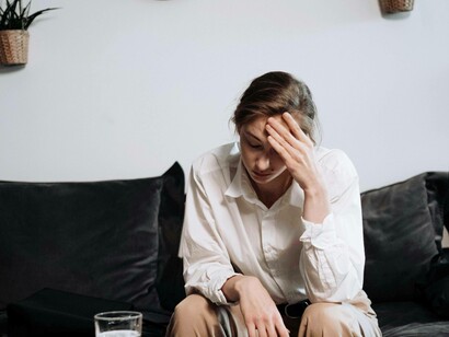 Una donna seduta su un divano, si appoggia la mano sulla fronte in un gesto di preoccupazione e stanchezza. Il suo sguardo pensieroso e l'ambiente circostante suggeriscono un senso di carico mentale o di frustrazione