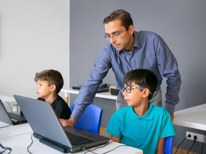 Teacher in glasses standing behind pupil and checking task on laptop