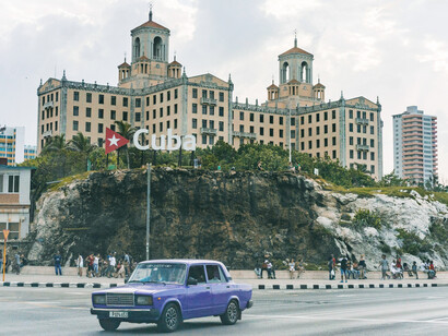 A purple sedan driving on the road near a building during the day, captured from El Malecón in Havana, Cuba