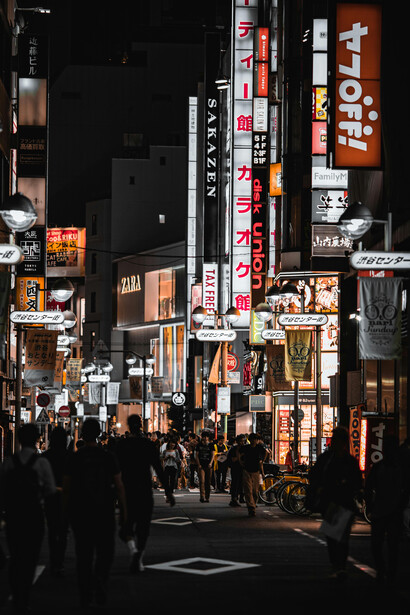 People walking along a city street lined with buildings and glowing street signs at night in Tokyo, Japan