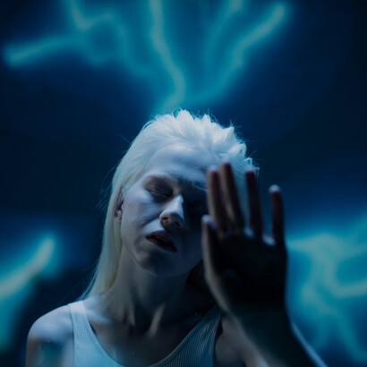 In a dimly lit room, a woman in white undergarments looks into the distance, her figure gently highlighted by streaming light rays