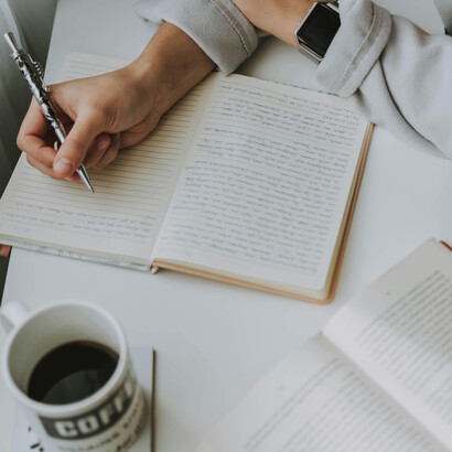 A writer pens stories in a notebook, with an open book and pen resting nearby