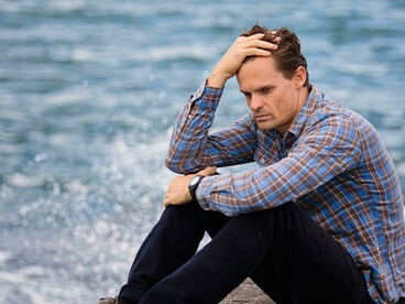 Wearing a blue and brown plaid shirt, the man touches his hair while appearing depressed and alone