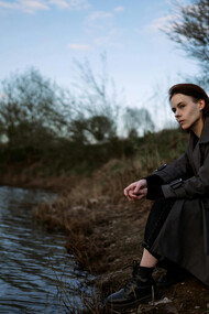 A young woman in a trenchcoat, contemplative, sitting at the shore of a small lake