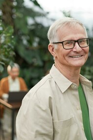 An elderly man in glasses smiling in a garden with another person in the background 