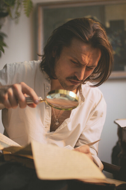 A solitary writer sits at a desk, smoking a cigarette and holding a sheet of paper in a dimly lit room