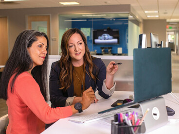Two businesswomen discussing sales strategies in an office, sitting at a desk with a laptop in front of them