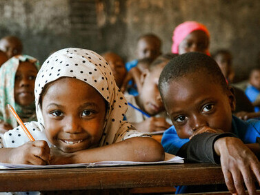 A smiling girl writes with a pencil in her classroom in Africa, with other students in the background, highlighting childhood education and academic focus