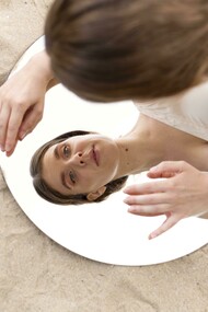Una donna si china verso uno specchio rotondo sulla sabbia, riflettendo il suo viso