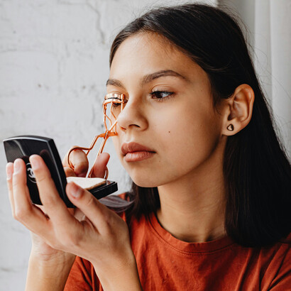 A young woman carefully curls her eyelashes as she finishes her makeup routine