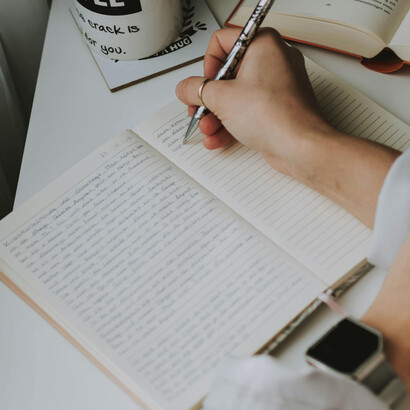 An author sits writing by hand in a notebook, surrounded by an open book and a pen