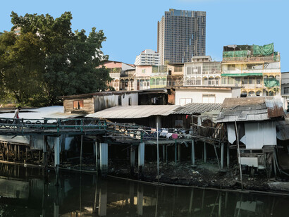 Slums line the riverbank while modern city buildings rise in the distance, illustrating the stark divide between rich and poor