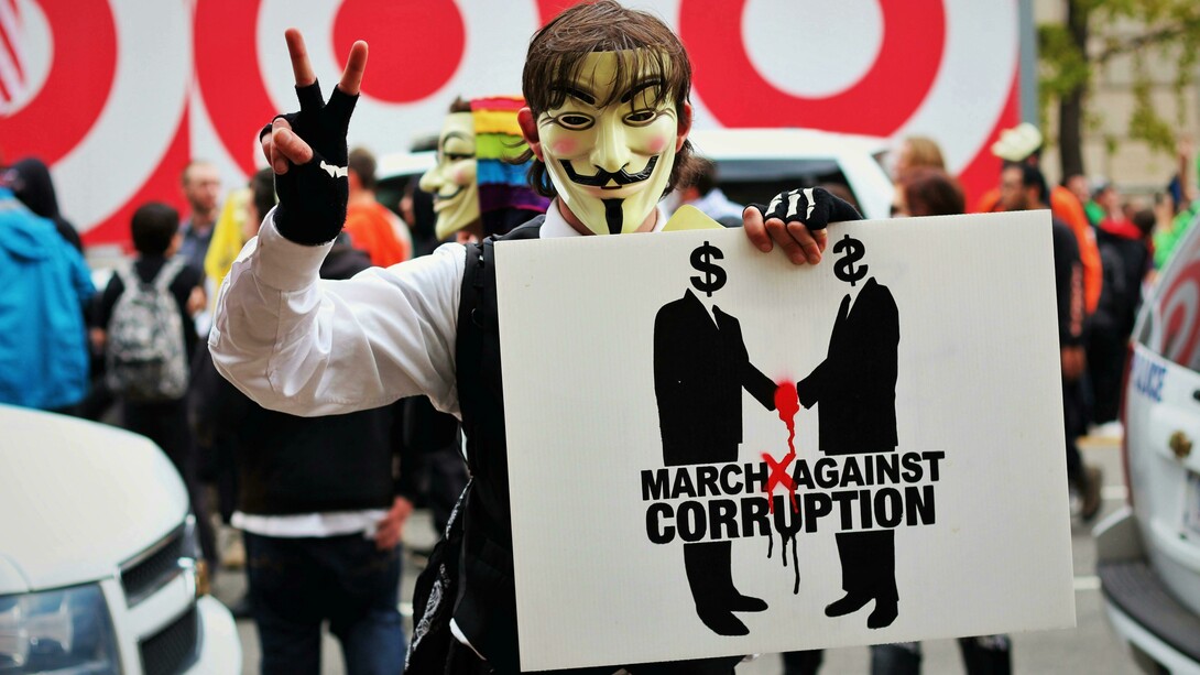 Person wearing a Guy Fawkes mask and holding a sign that reads “March Against Corruption”