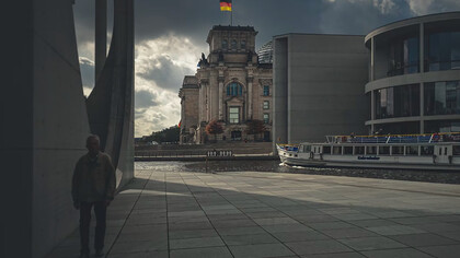 An elderly man walks along a gray concrete path beside a brown building in Berlin, Germany
