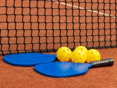 Yellow pickleballs nestled between two blue paddles on a clay-colored court highlights the accessibility of pickleball, a sport that not only promotes cardiovascular health and agility but also fosters community and social interaction
