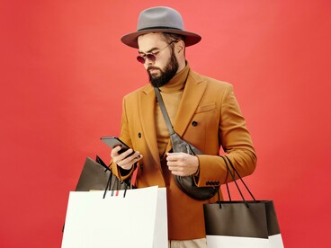A man in a brown blazer walking with shopping bags and checking his phone, symbolizing how consumerism intersects with the search for connection and identity