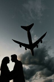 Silhouettes of a couple sharing a farewell embrace under a plane flying across the sky at the airport