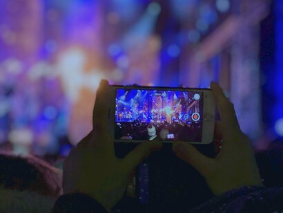Apresentação gravada no celular. Grande parte mantinha os olhos fixos na tela, como se estivessem em outra dimensão — registrando o instante para o futuro, mas ausentes do agora. De onde eu estava, os celulares erguidos lembravam escudos luminosos, como barreiras entre o real e o possível