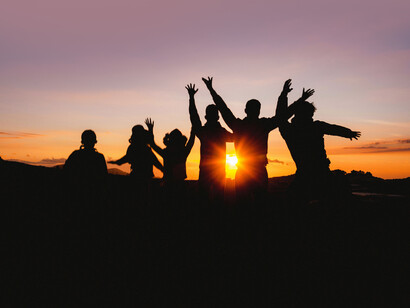 People, seen in silhouette, raise their hands in joy against a golden-hour sunset