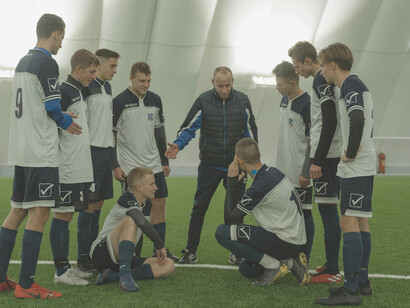 A football coach addressing his players while the director of football and manager confer nearby