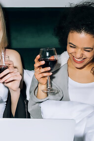 Two women with wine in hand looking at computer screen on bed and smiling